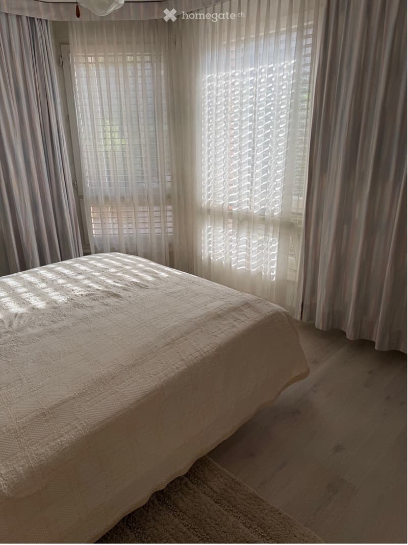 A well-lit bedroom with a neatly made bed covered in a textured beige duvet, positioned near large windows with sheer white curtains allowing natural light to filter in.
