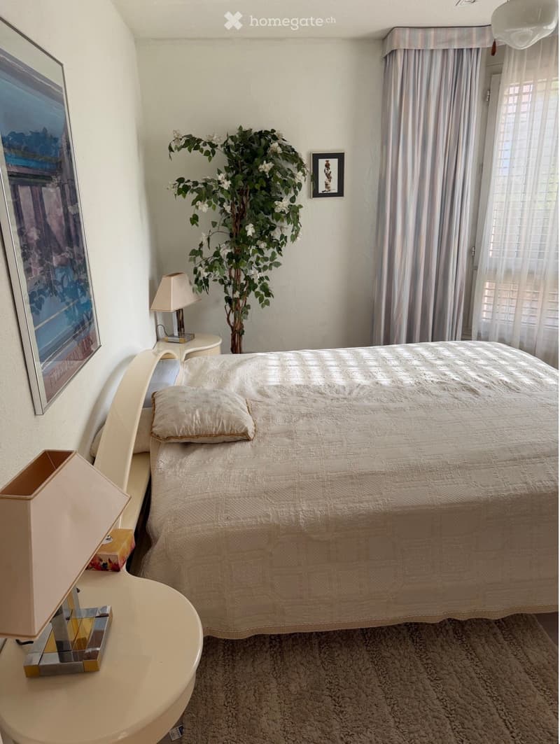 Cozy bedroom with a neatly made bed, nightstand, lamp, and decorative plant. Features a large window with blinds and artwork on the walls.