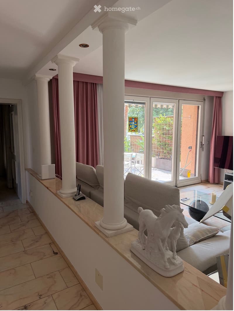 Living room with a sofa, a glass coffee table, a decorative statue, and a candle on the table. The room features large windows and a balcony with outdoor furniture visible.