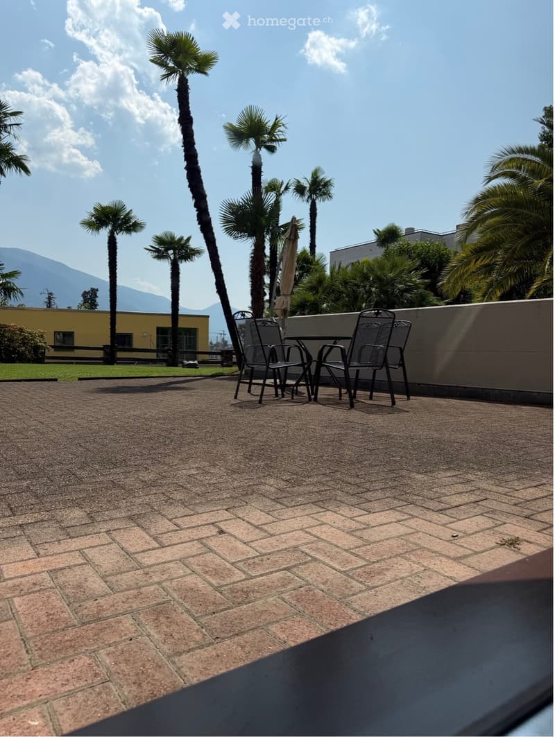 Brick-paved balcony with a small table and two chairs, surrounded by palm trees and a distant mountain view.