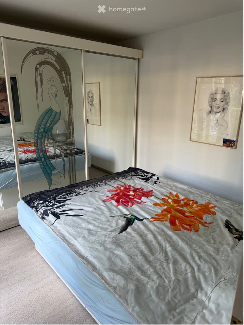 A bedroom with a large bed covered in a decorative comforter featuring floral and bird designs. A large mirror with an ornate frame is visible, along with a nightstand and lamp. Artwork is displayed on the walls.