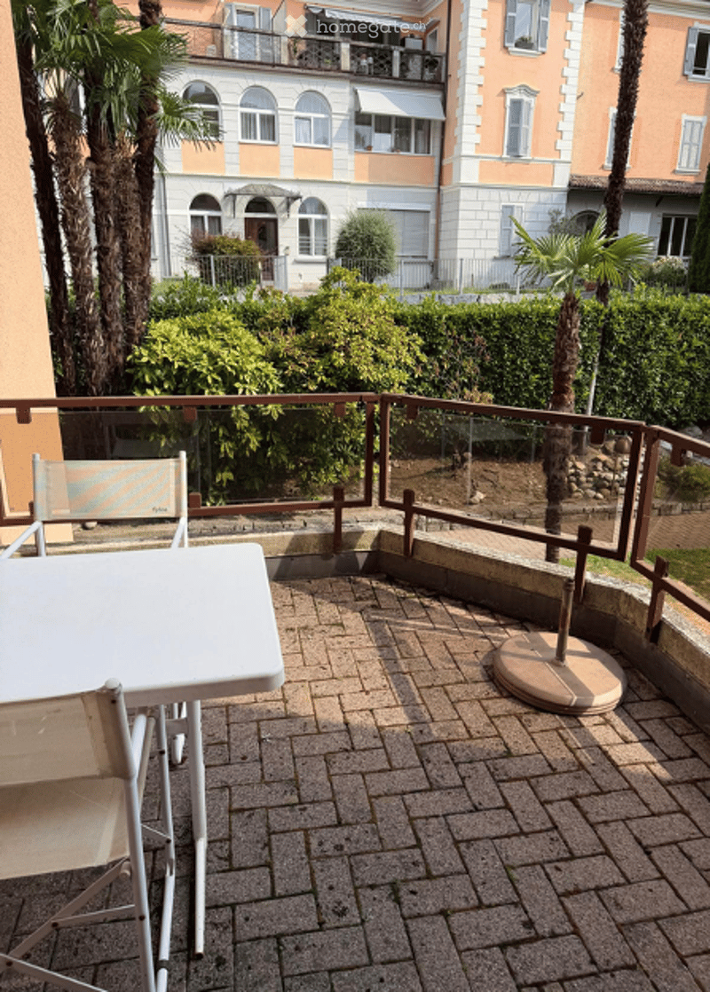 Balcony with a white table and chairs, surrounded by greenery and palm trees, overlooking a residential area.