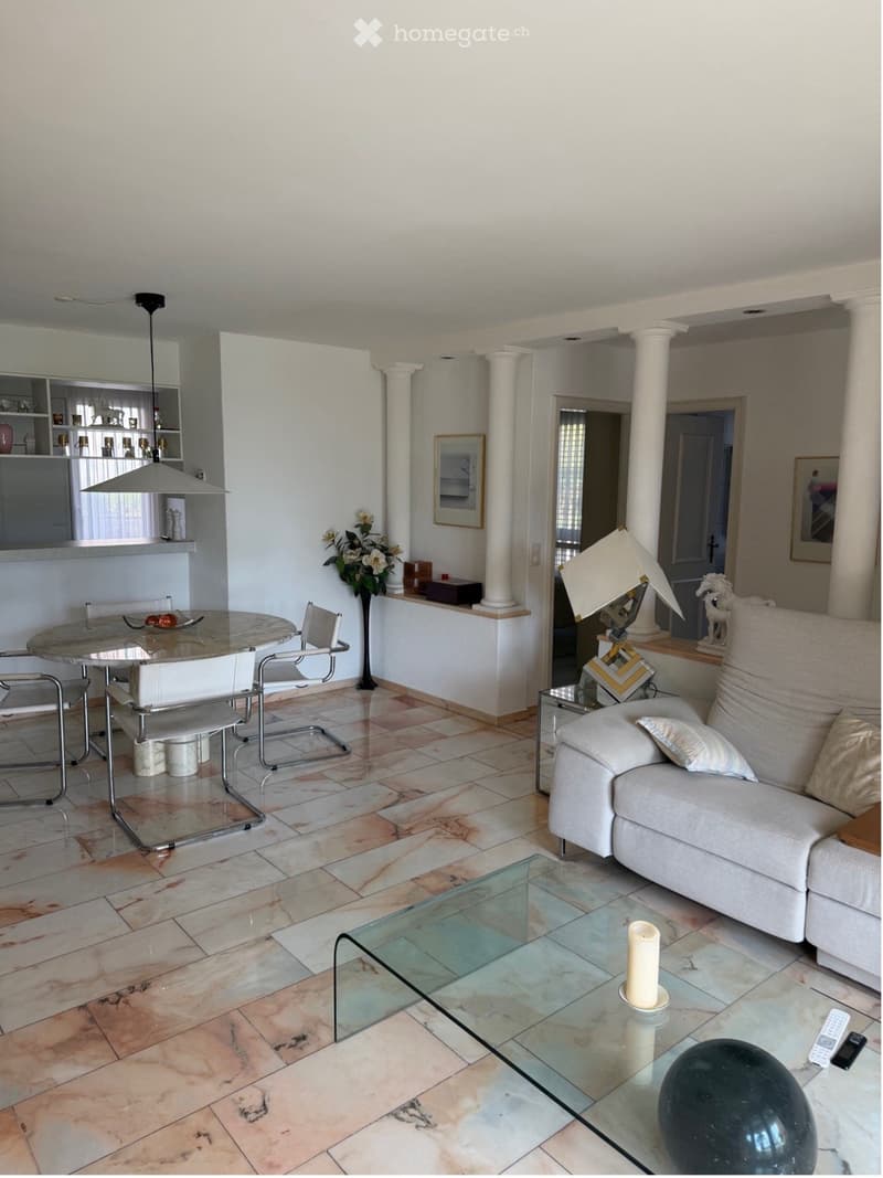 Living room with a white sofa, glass coffee table, dining table with chairs, decorative lamp, vase with flowers, and picture frame on the wall.