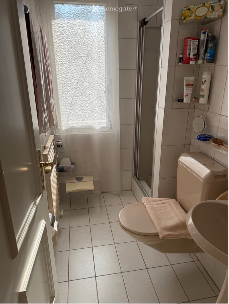 Bathroom with white tiled floor and walls, toilet with a towel on the seat, sink, shower with a curtain, radiator, and shelves with toiletries.