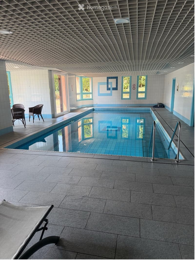 Indoor swimming pool area with tiled walls, large windows, and two chairs near the pool.