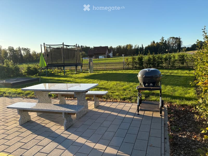 Outdoor patio with stone table, benches, grill, and umbrella, surrounded by a grassy lawn and trees.