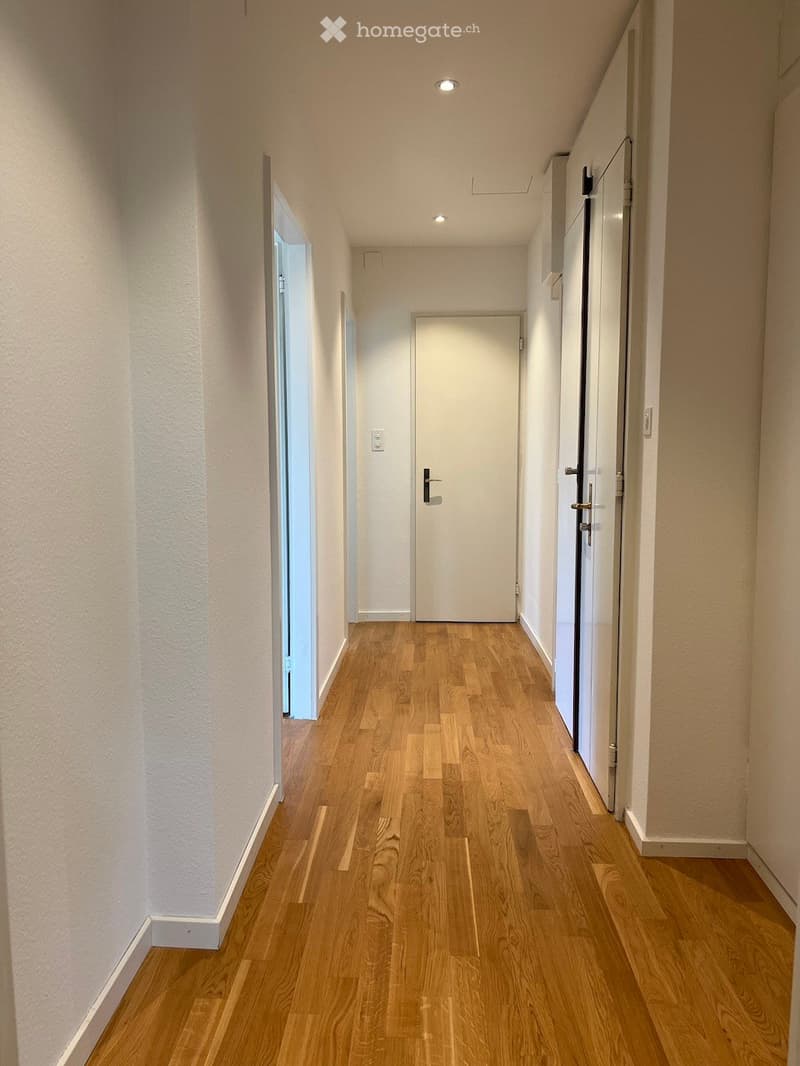 A narrow hallway with light wooden flooring, white walls, and recessed ceiling lights. There are two doors visible, one on each side, and a small open doorway leading to another area.