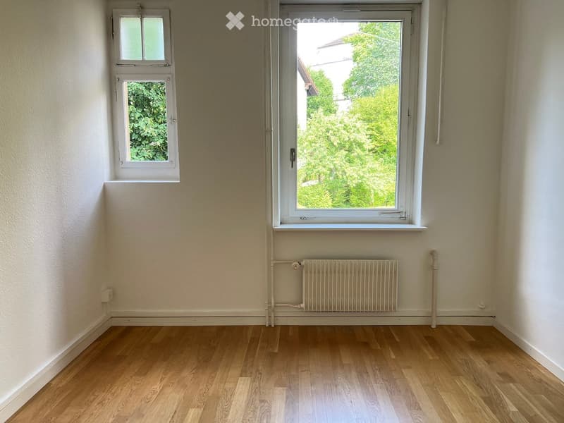 Empty room with wooden flooring, white walls, and two windows showing greenery outside. A radiator is mounted on the wall below the larger window.