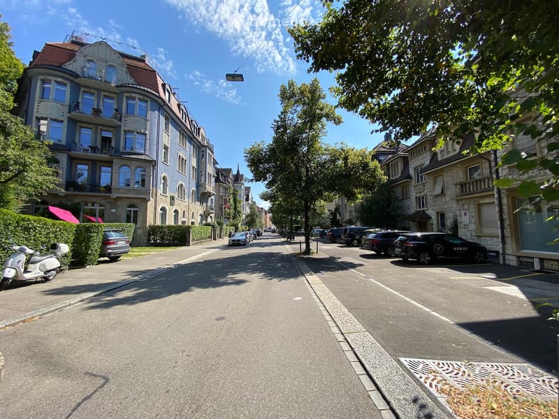 Street view of an avenue lined with apartment buildings and parked cars with green spaces and streetlights.