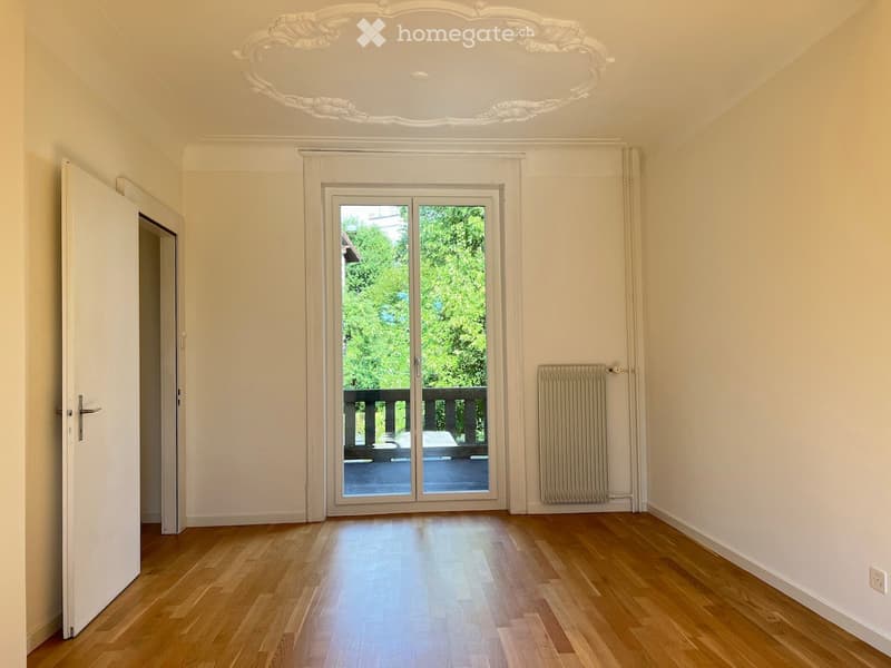 Empty room with wooden flooring, white walls, large sliding glass door leading to a balcony, radiator, and decorative ceiling molding.