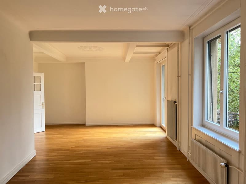 Empty room with wooden flooring, white walls, and large windows allowing natural light. The space features a radiator under one window and a door leading to another room.