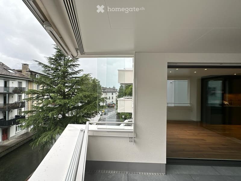 Modern balcony with large glass windows, wooden flooring, and a view of a tree-lined street and neighboring buildings.