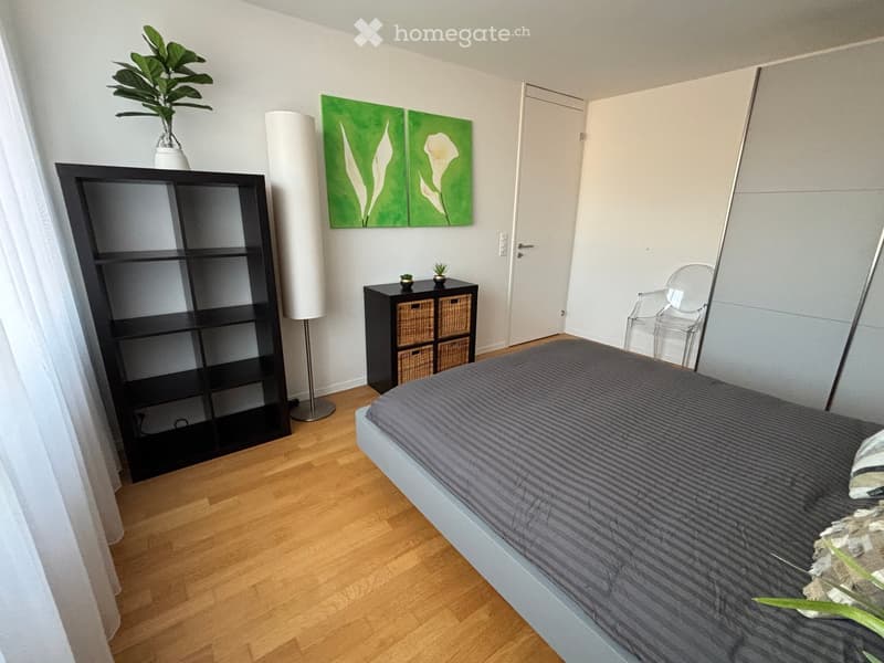 Modern bedroom with a grey bed, black shelving unit, floor lamp, wicker storage unit, and a clear plastic chair. The room features wooden flooring and a large green artwork on the wall.