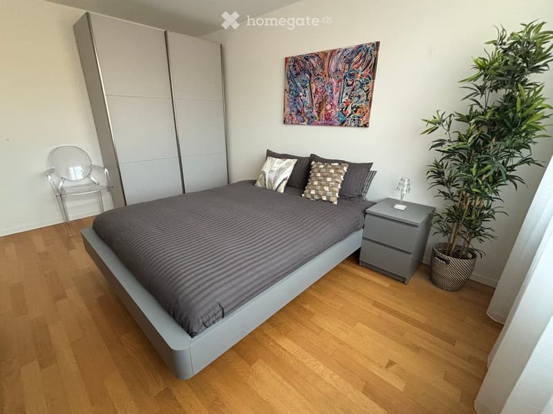 Modern bedroom with a grey duvet-covered bed, nightstand, decorative pillows, a sleek chair, and a potted plant near the wall.
