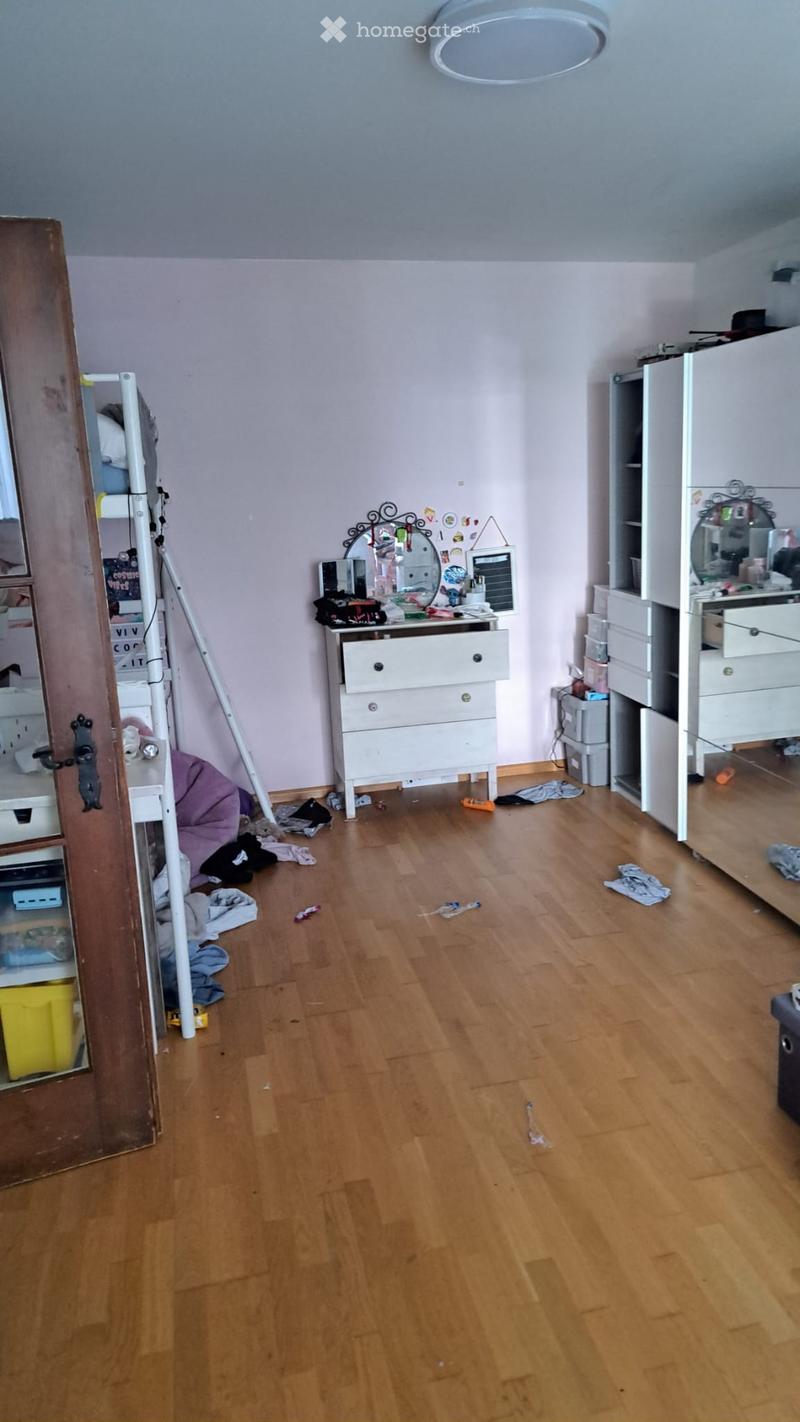 Empty room with wooden flooring, light-colored walls, and scattered items including a ladder, a dresser, and a mirror. The room appears to be in the process of being organized or cleaned.