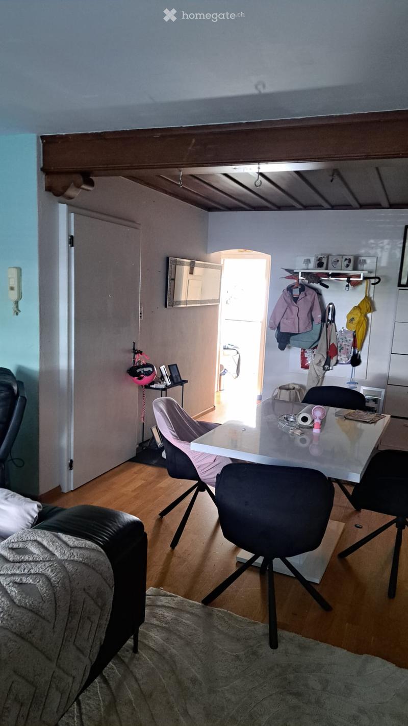 A cozy living room with a black leather sofa, a white coffee table, a pink armchair, a coat rack, and a small table with a laptop. The room has wooden flooring and a slanted ceiling.