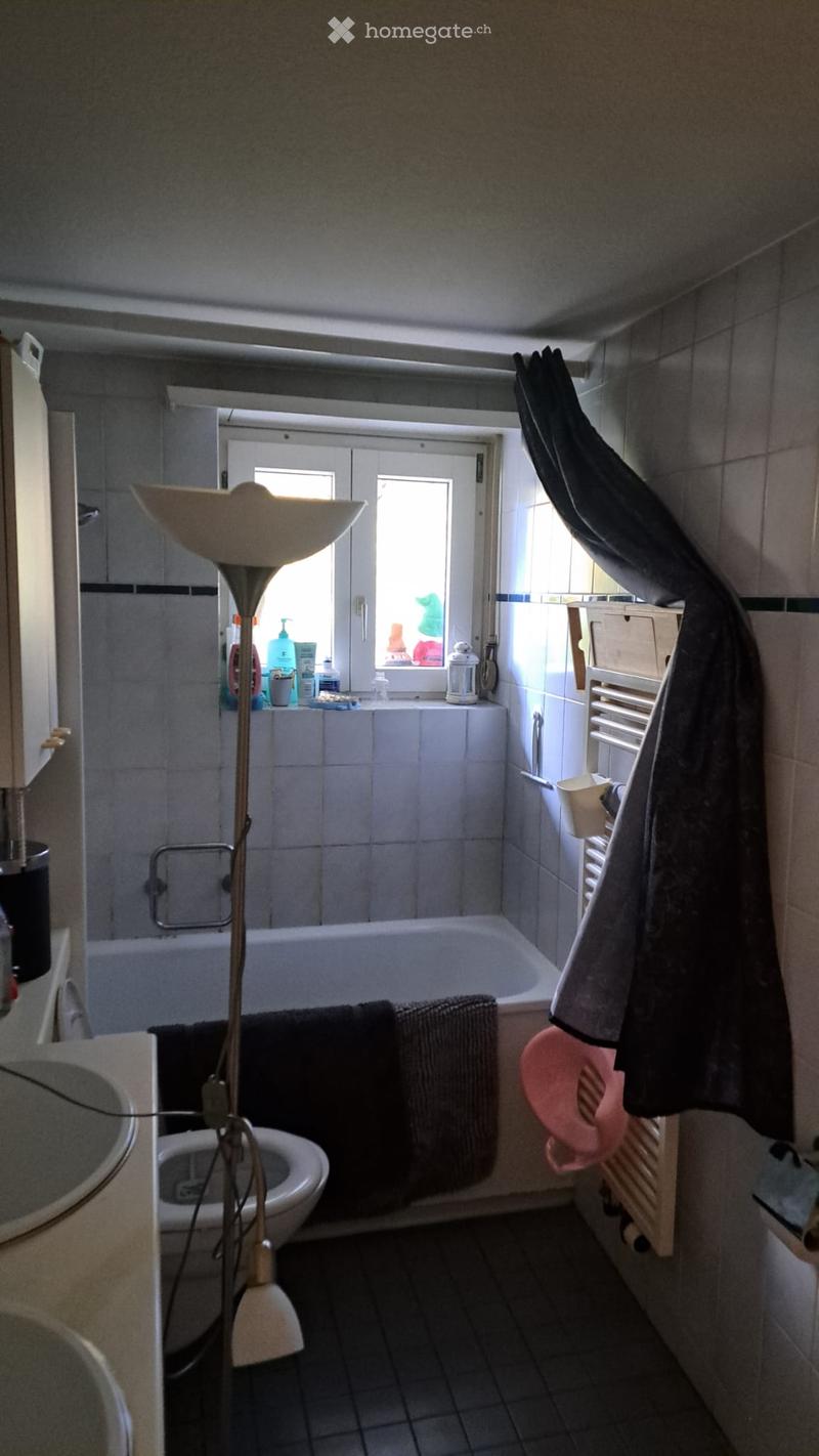 Bathroom with a toilet, bathtub, sink, towel rack, floor lamp, and a window with a shower curtain. Towels are placed on the edge of the bathtub and on the towel rack.
