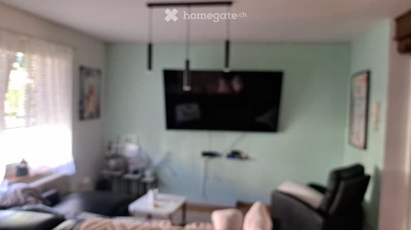 A modern living room with a sofa, a small table, a chair, a television mounted on the wall, and picture frames. The room has light green walls and a large window with white curtains.