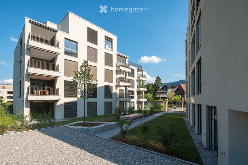 Modern multi-story residential building with white facade, balconies, and contemporary design. The courtyard features a small tree, seating area, and paved pathways.