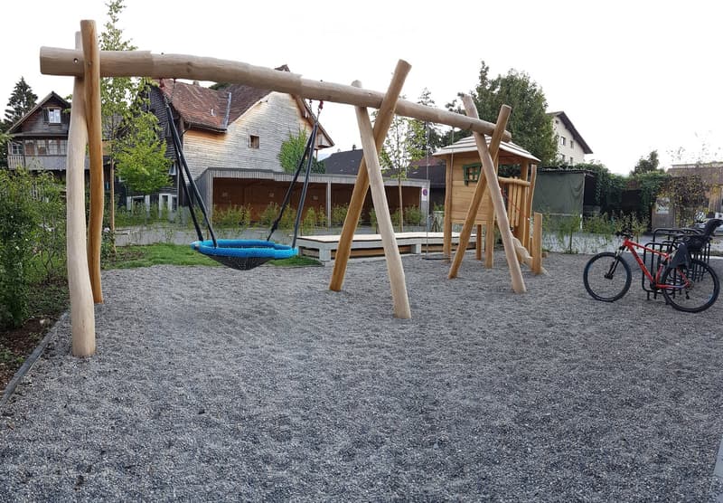 Playground area with a blue nest swing, wooden climbing frame, and a red bicycle parked nearby. The ground is covered with gravel, and there are wooden benches and a small wooden playhouse in the background.