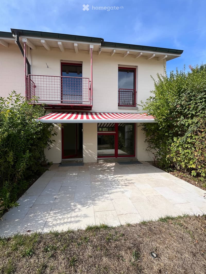 Two-story building with white brick facade, red-framed windows and doors, a striped awning, and a small balcony with a metal railing.