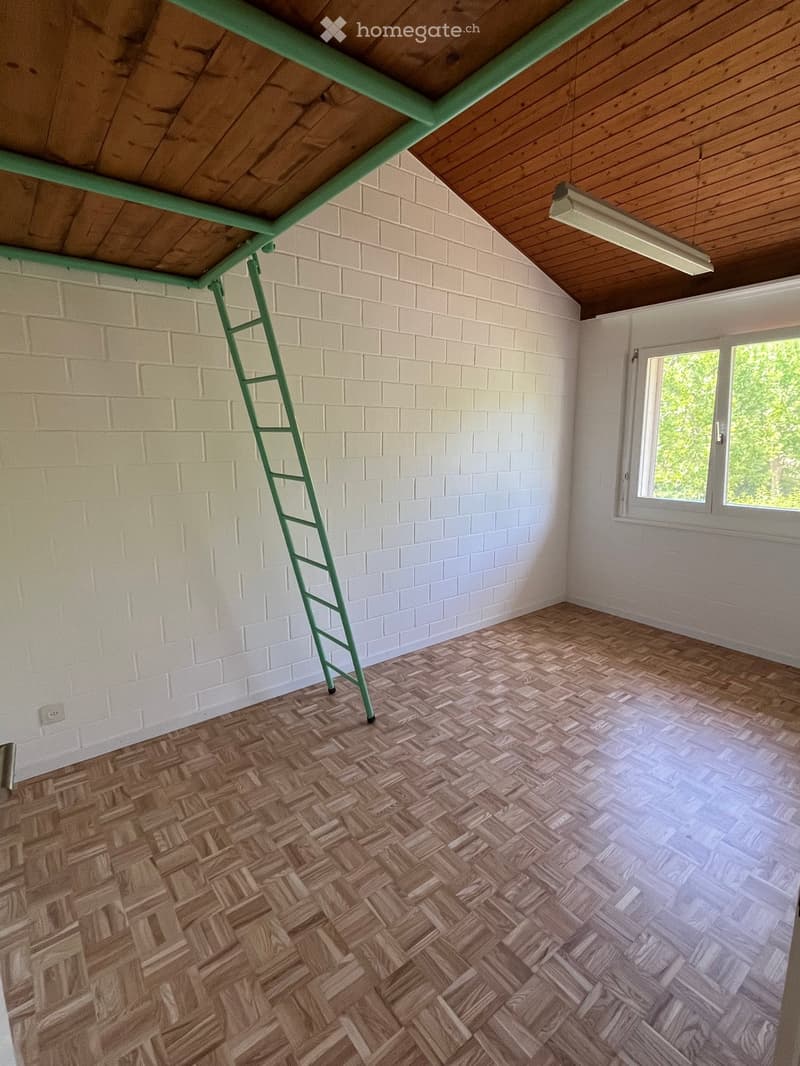 Empty room with wooden herringbone flooring, white brick walls, and a green ladder leaning against the wall. A window with white frames allows natural light to enter.