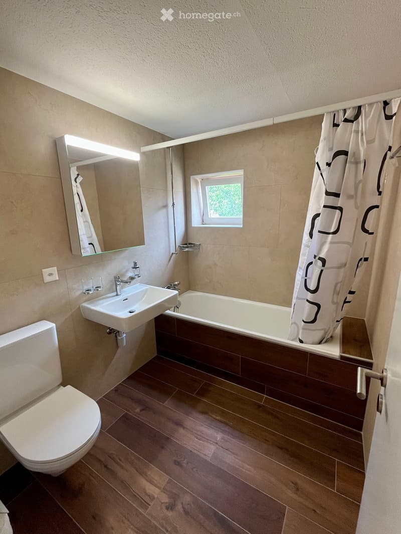 Modern bathroom with a toilet, sink, bathtub, mirror, and a window. The floor is wooden, and the walls are tiled.