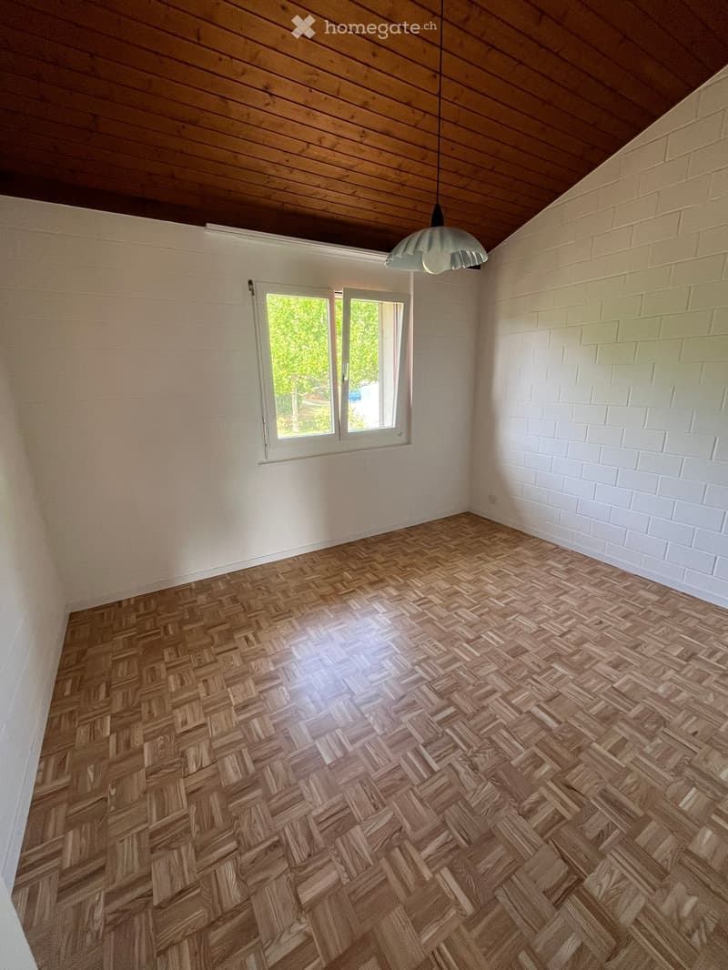 Empty room with wooden herringbone flooring, white brick walls, and a large window with white frame. A single hanging light fixture is visible.