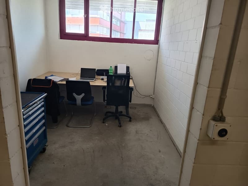 Office space with a desk, laptop, chairs, a bottle, and a cabinet. The room has white brick walls and a large window with blinds.