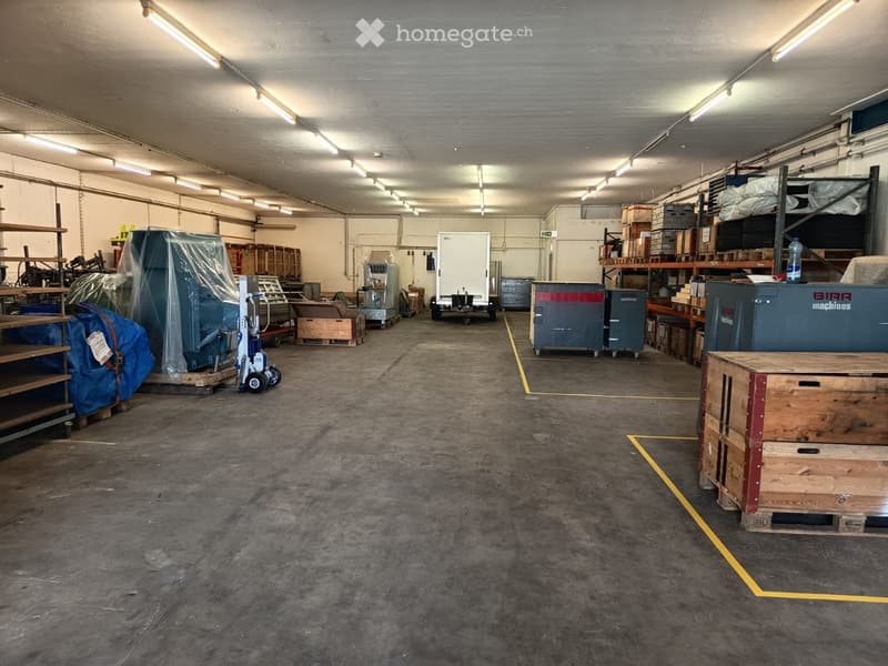 Large, empty warehouse space with concrete floors, exposed ceiling beams, and scattered storage items including crates, covered machinery, and shelving units.