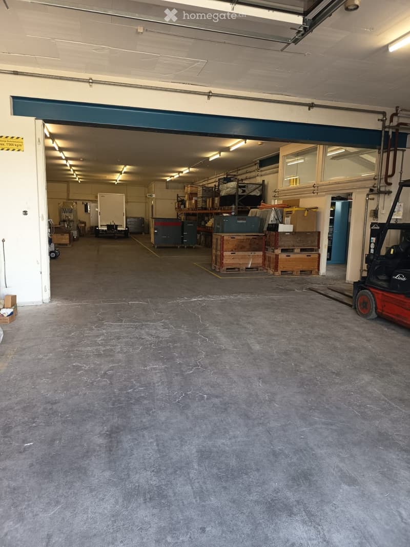 Empty warehouse space with concrete floor, large open doorway, and some stacked crates and equipment visible in the background.