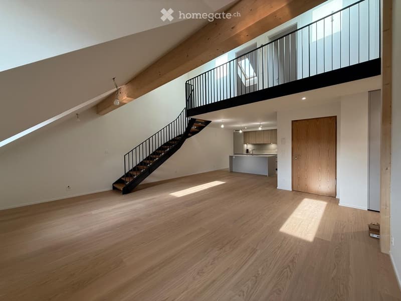 Open-concept unfurnished room with wooden flooring, white walls, and a staircase leading to an upper level. Features include a modern kitchen area with cabinets and a countertop, and a balcony with a metal railing visible in the background.