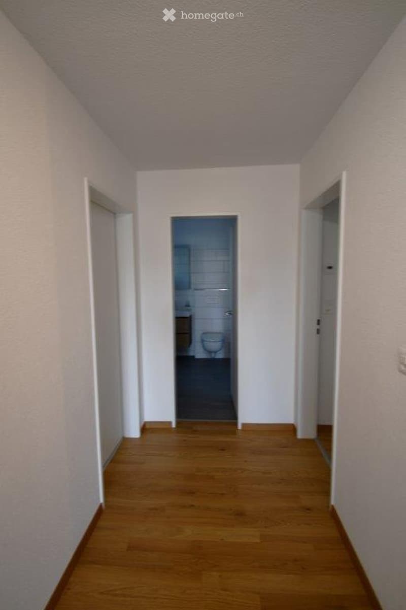 A narrow hallway with wooden flooring and white walls, leading to a partially visible bathroom with a toilet and sink.