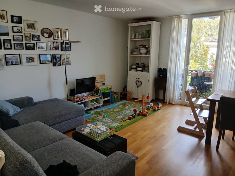 Living room with a grey sectional sofa, coffee table with books, a play mat with toys, a TV, a shelf with dishes, and a dining table with chairs. The room has wooden flooring and large windows with white curtains.