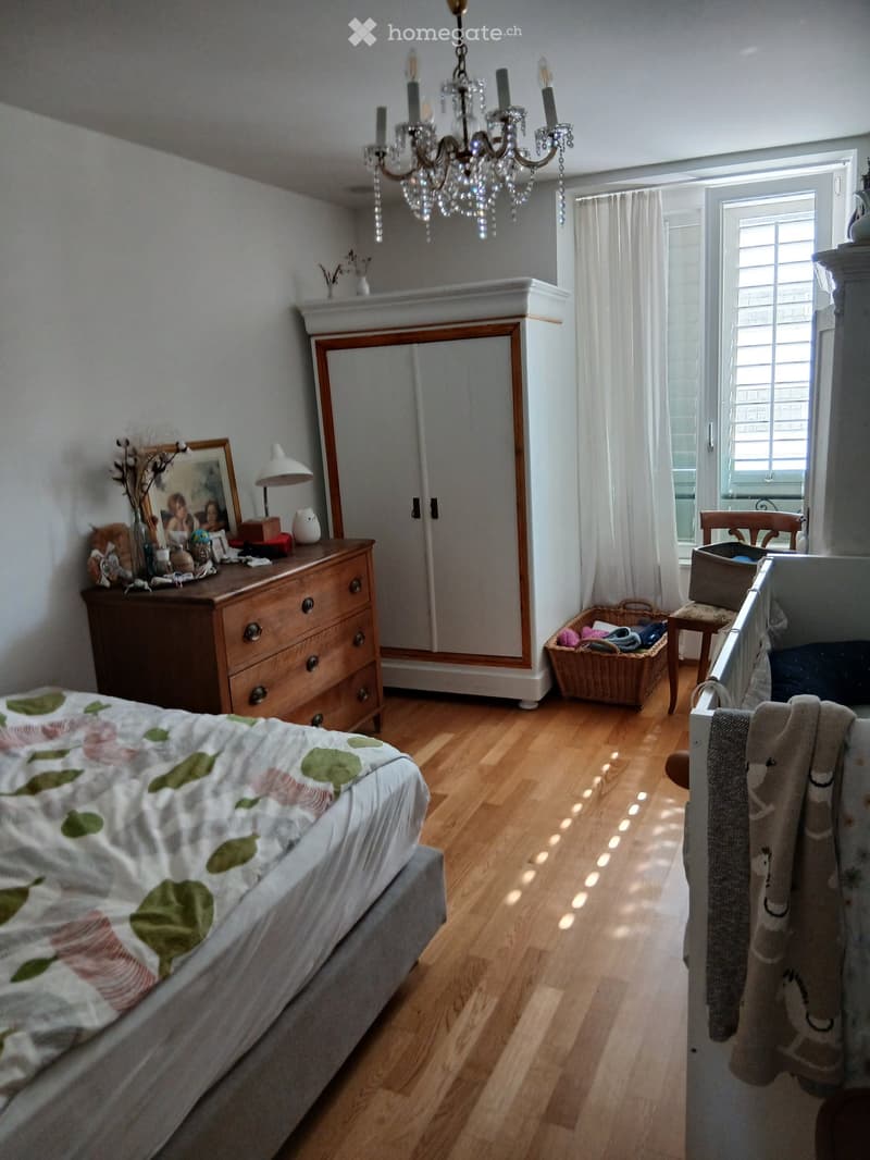 Well-lit bedroom with wooden flooring, a bed with a floral quilt, a dresser with a mirror, a wardrobe, a wicker basket, and a small table with a chair near the window.