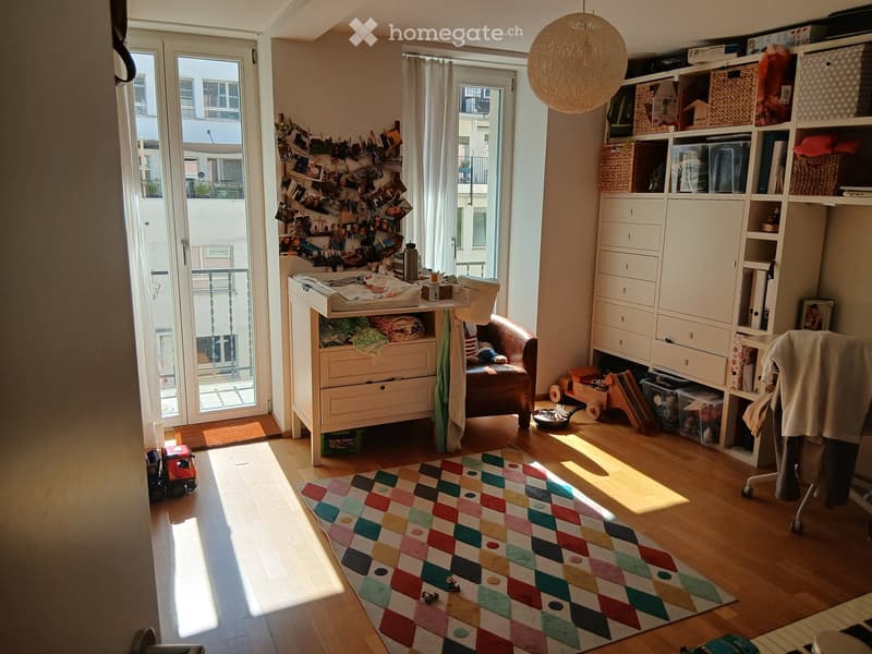 Brightly lit room with wooden flooring, large windows, and a balcony. Features a colorful geometric rug, a leather armchair, a small table with baby items, a shelf with books and toys, and a large round pendant light.