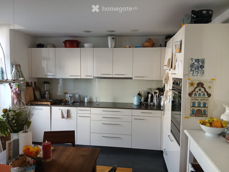 Modern kitchen with white cabinets, stainless steel appliances, and a wooden dining table with chairs. The counter features a sink, blender, and various kitchen items.