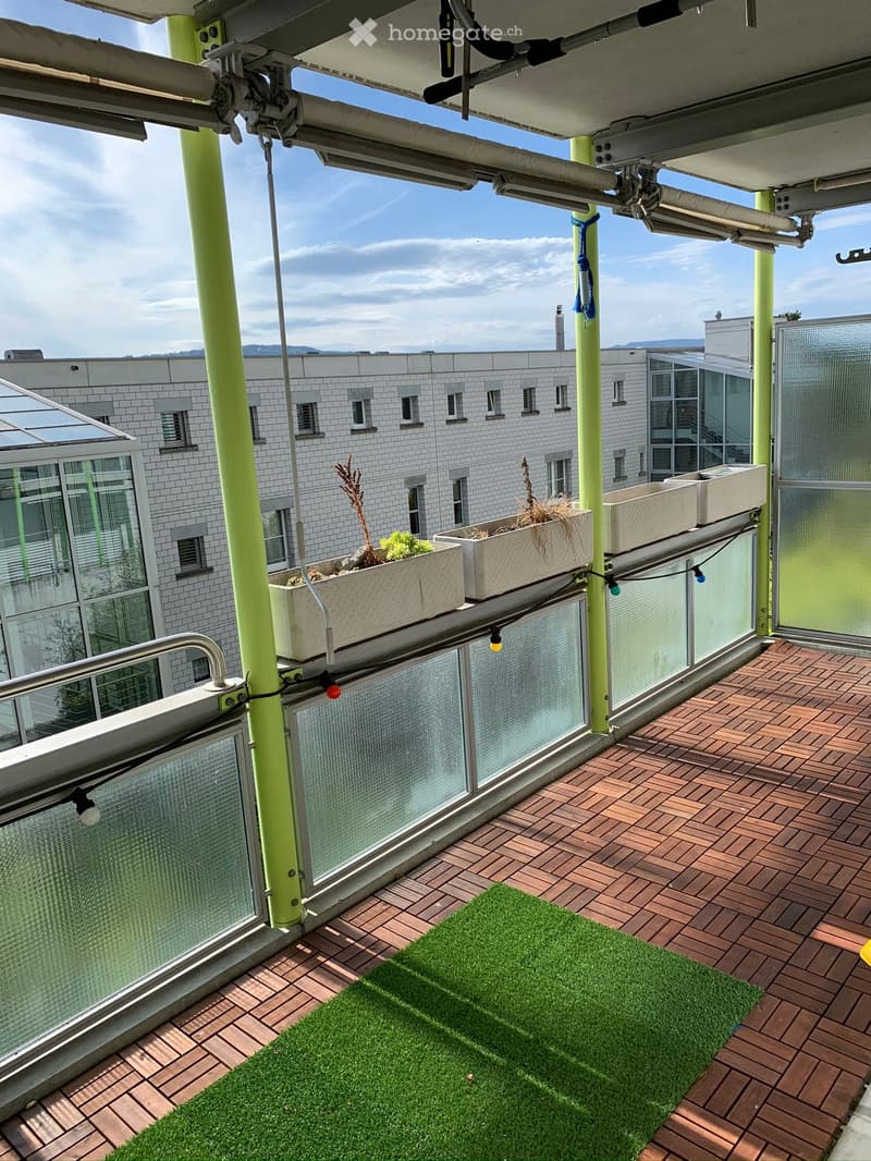 Balcony with wooden decking, green artificial grass, and several white planters with plants. The balcony has glass railings and is equipped with small lights.