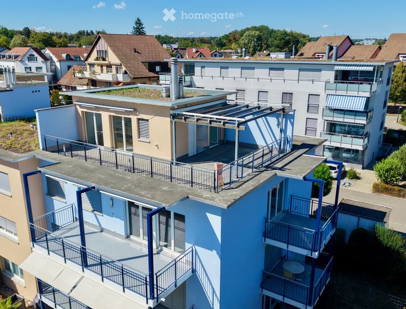Aerial view of a modern multi-story residential building with balconies, surrounded by other houses and greenery.