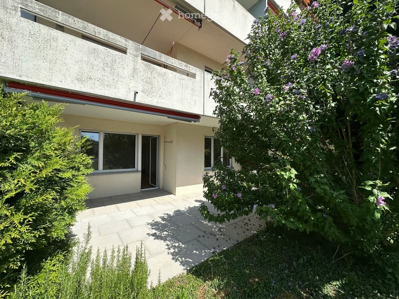 A modern house with a balcony, surrounded by lush greenery and flowering bushes. The entrance features a tiled patio area.