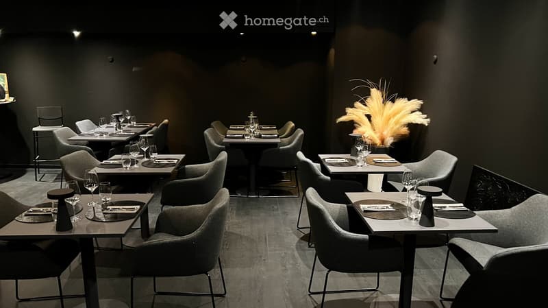 Modern restaurant interior with multiple tables and chairs, wine glasses, plates, and cutlery set for dining. A decorative flower arrangement is placed on a table in the corner.