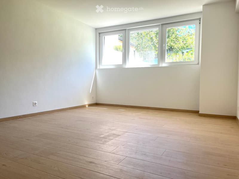 Empty room with light wooden flooring and white walls, featuring large windows with white frames and visible greenery outside.