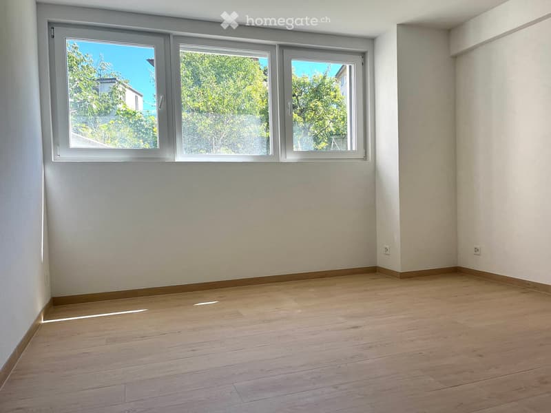Empty room with wooden flooring, white walls, and large windows with a view of greenery outside.