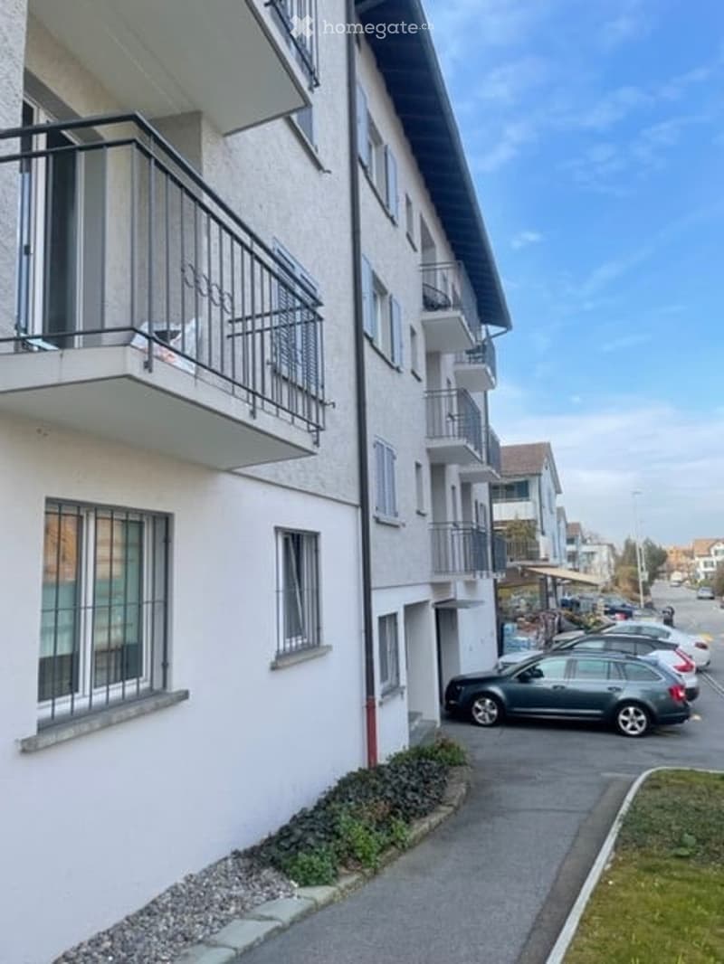 Modern multi-story residential building with white facade, multiple balconies, and parked cars in the driveway.