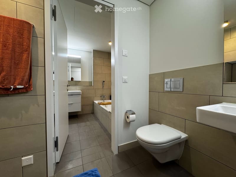 Modern bathroom with a toilet, sink, bathtub, and mirror. Features tiled walls and floor, a towel on the side, and a small wooden tray in the bathtub.