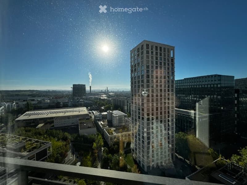 A modern high-rise building with reflective glass windows, surrounded by other structures and greenery, under a clear blue sky with the sun shining brightly.