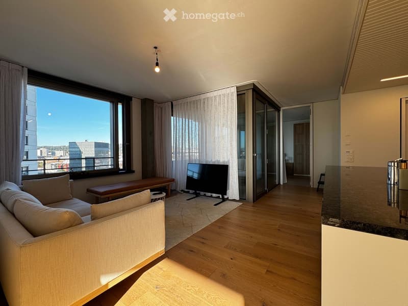 Modern living room with large windows offering a city view, a cream-colored sofa, a wooden bench, and a flat-screen TV on a stand.