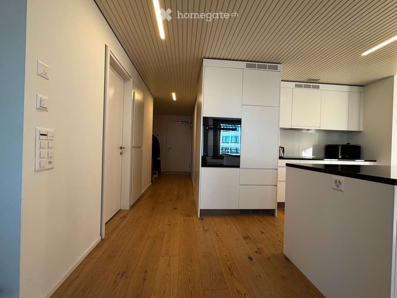 A modern hallway with wooden flooring, white walls, and a small kitchen area visible at the end. The kitchen features white cabinets, a countertop, and a microwave.