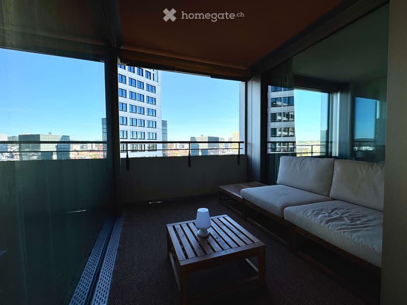 Balcony with a modern design, featuring a white sofa, wooden coffee table, and table lamp, overlooking a cityscape with tall buildings.
