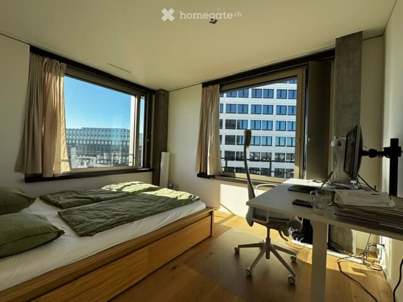 Bright bedroom with large windows offering a city view, featuring a wooden bed with green bedding, a desk with a computer setup, and a chair.